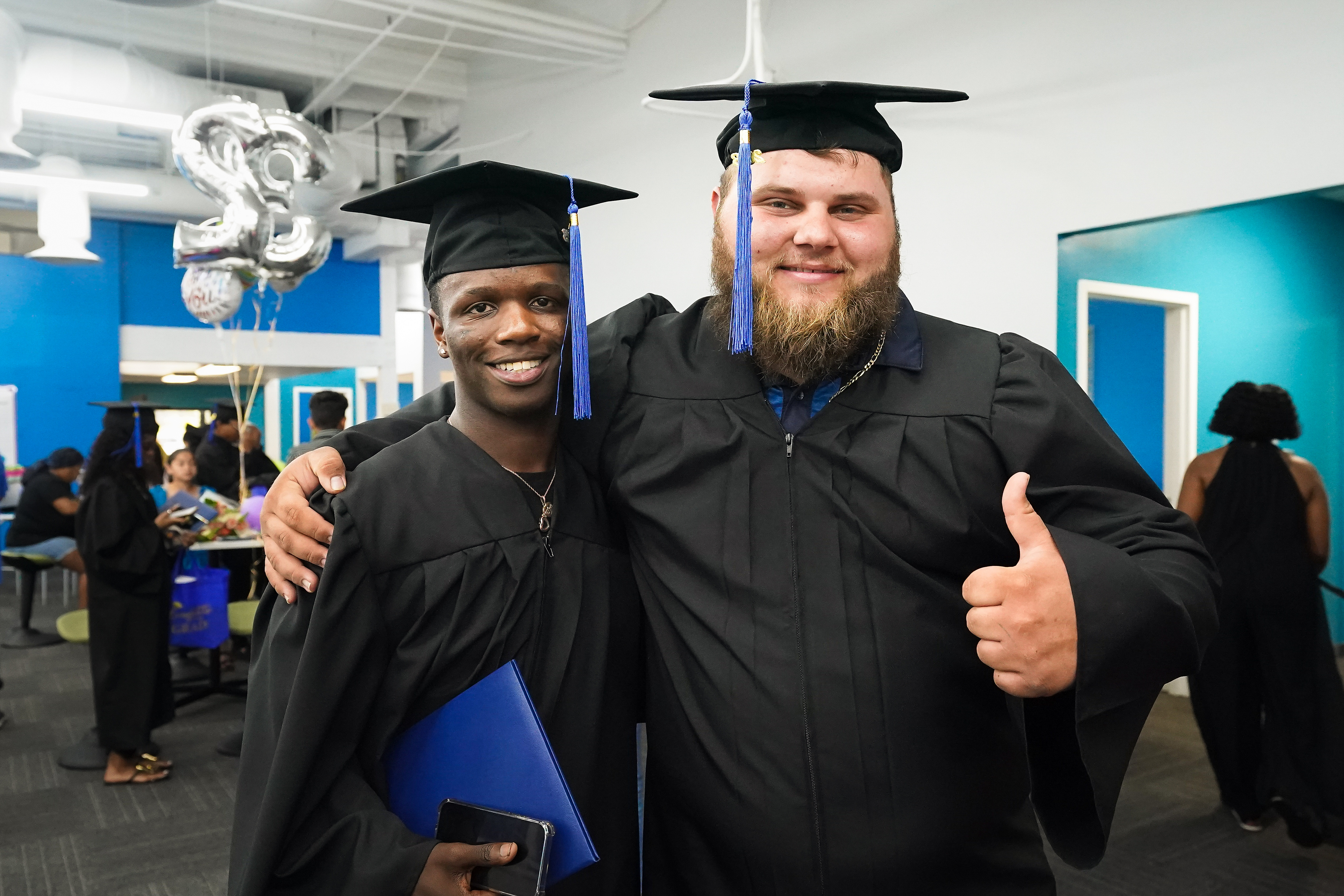 Two male graduates stand next to each other, one gives a thumbs up.