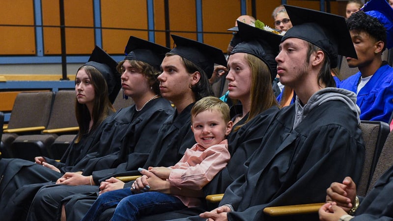 Young boy smiles on mom's lap as she sits in an auditorium during graduation ceremony