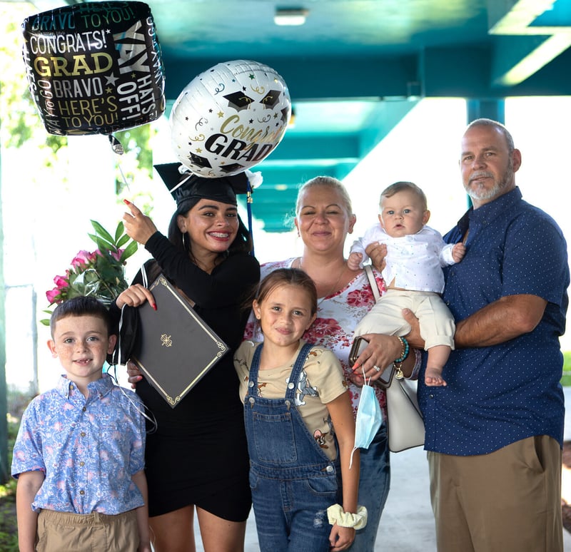 Female graduate holds diploma and balloons, smiling surrounded by family