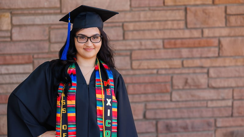 Female graduate smiles in cap and gown in front of a brick wall.