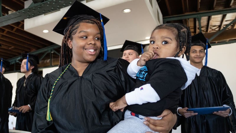 Young mom and high school graduate Tamia smiles at graduation ceremony holding her baby